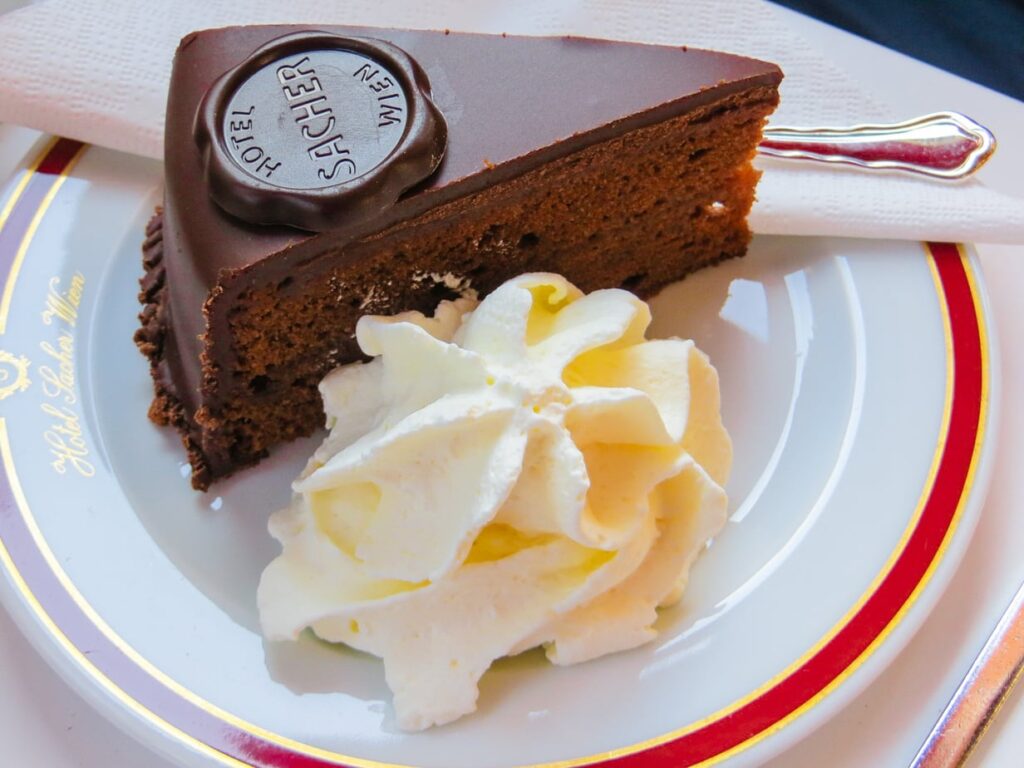 A slice of Viennese Sachertorte with glossy chocolate icing, a layer of apricot jam, and whipped cream on the side.