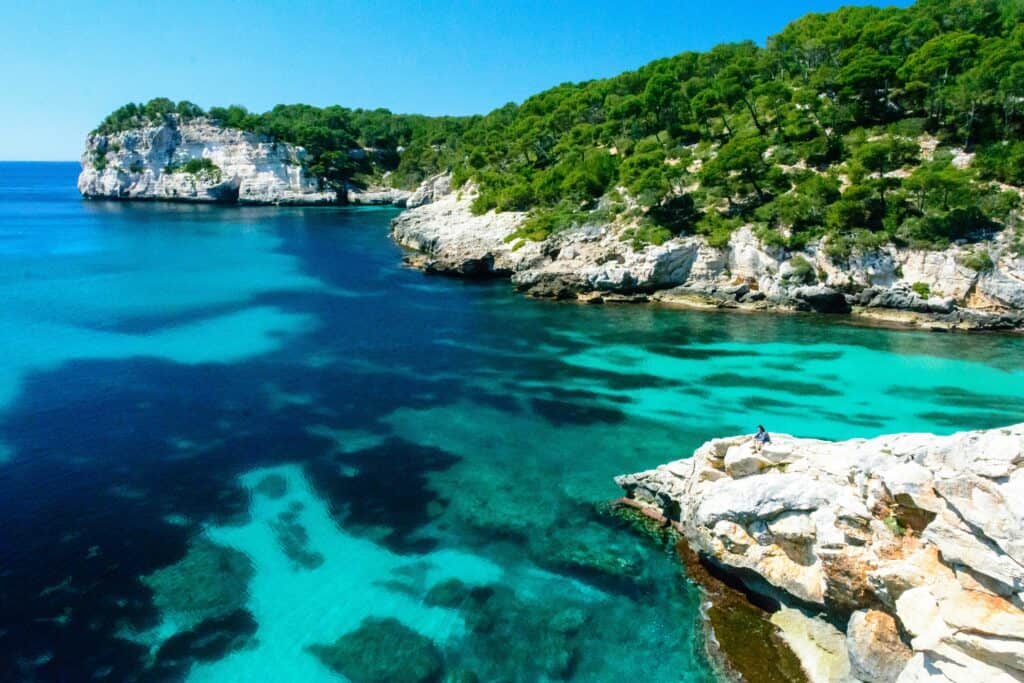 Cala Mitjana beach in Menorca, Spain, with turquoise water, white sand and pine-covered cliffs surrounding the secluded cove
