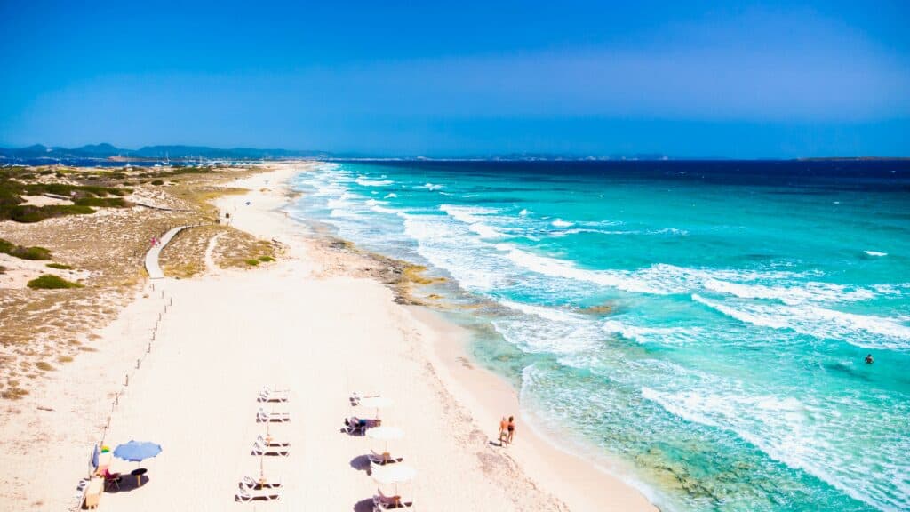 Formentera beach in Spain with white sand, clear turquoise water and people relaxing along the shoreline.
