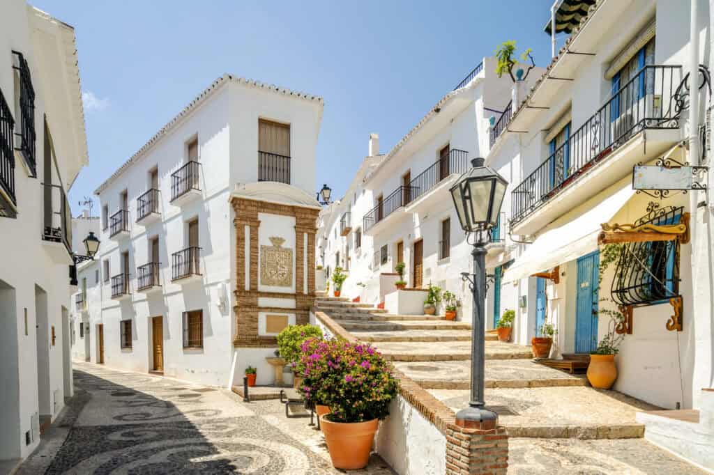 “Narrow whitewashed street in Frigiliana, Spain, with cobblestones, blue doors and flower pots filled with bright geraniums.