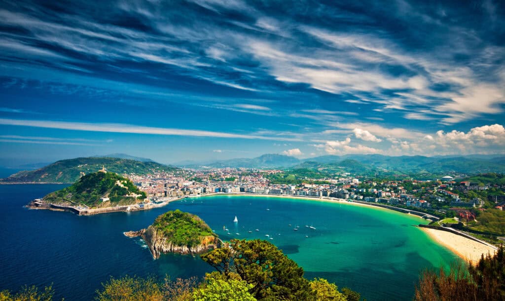 Panoramic view of La Concha Bay in San Sebastián, Spain, with golden beach, turquoise water and green hills in the background.”