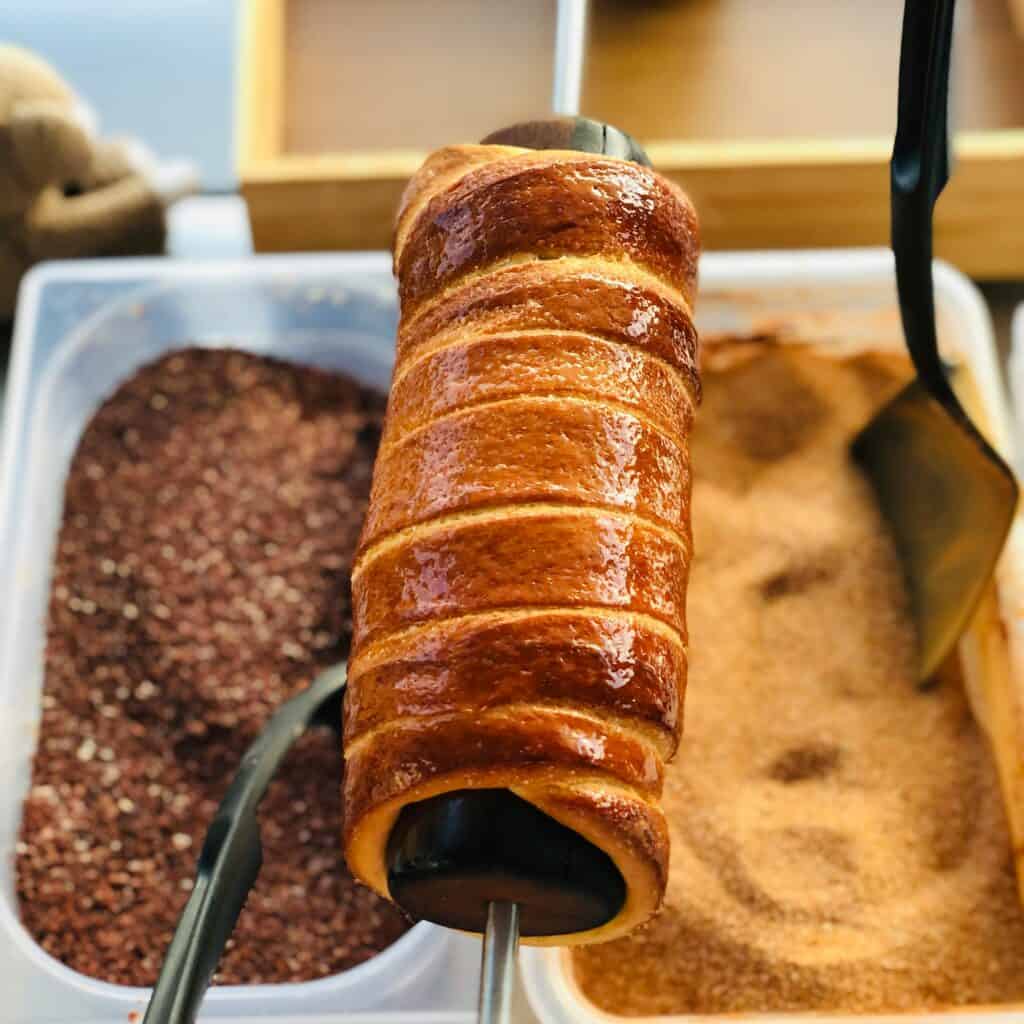 Fresh Hungarian chimney cake (kürtőskalács) baking on a rotating spit, coated in sugar and caramelising to a golden brown crust.
